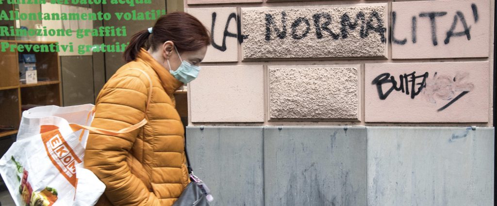 A woman wearing a protective mask walks next an inscription painted on  the wall of a building reading in italian "normality", Genoa, Italy, 21 April 2020. Italian Prime Minister Giuseppe Conte, during his speech to the Chambers, said that the masks and the social security distancing will be maintained until a vaccine against Covid-19 wil not be discovered. ANSA/LUCA ZENNARO